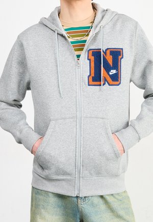 Personne portant un sweat à capuche gris à fermeture éclair avec un patch en forme de lettre N bleu et orange, les mains dans les poches devant un t-shirt rayé et un jean clair délavé.