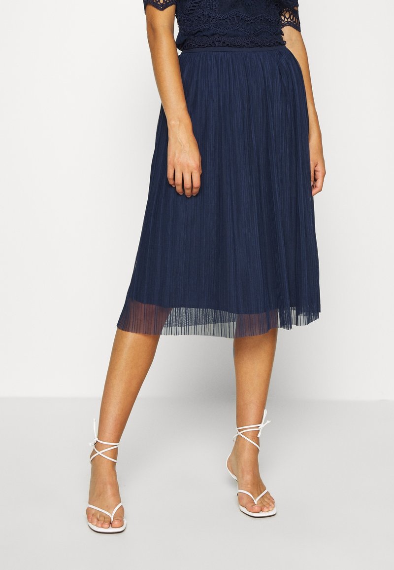 Jupe midi plissée bleu marine avec une texture légère plissée, taille froncée et ourlet transparent. Associée à des sandales blanches à lanières.