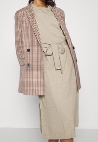 Blazer croisé à carreaux marron et crème porté sur une robe en maille beige avec une taille nouée. Tissu texturé et motifs contrastés.