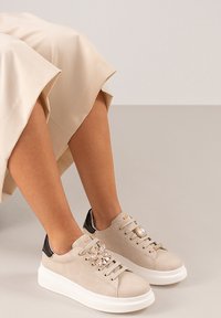 Zapatillas de ante beige con suela blanca, con detalles en negro y hardware decorativo en los cordones, parcialmente visibles debajo de una falda de color claro.