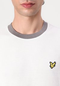 White short-sleeve shirt with a grey ribbed collar and a black-bordered yellow bird emblem on the chest. Textured fabric.