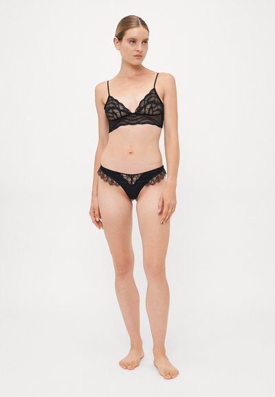 Woman standing barefoot wearing a black lace bralette with thin straps and matching lace-trimmed underwear against a plain white background.
