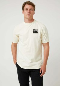 Camiseta de algodón color crema con mangas cortas, con una impresión negra llamativa que dice "SUEÑA EN GRANDE" junto a "ALTONADOCK".