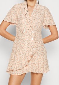 Robe à manches courtes, style portefeuille, en tissu pêche clair à motif floral. Dotée d'un ourlet à volants et de détails avec des boutons.