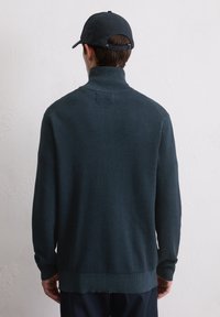 Dunkelgrüner Pullover mit hohem Kragen, gerippter Textur und langen Ärmeln. Auf der Rückseite minimalistische Details und ein gerader Saum.