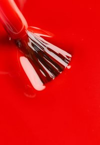 Esmalte de uñas rojo con un acabado brillante, con un pincel ancho sumergido en el líquido. La superficie parece lisa y reflectante.