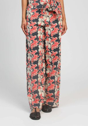 Pantalon à jambes larges avec motif floral rouge et beige sur fond noir, porté avec des chaussures plates tissées noires, mains reposant le long du corps.
