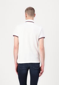 Vit polo med krage som har röda och marinblå ränder, korta ärmar och en figurnära design, bärs med mörka jeans.