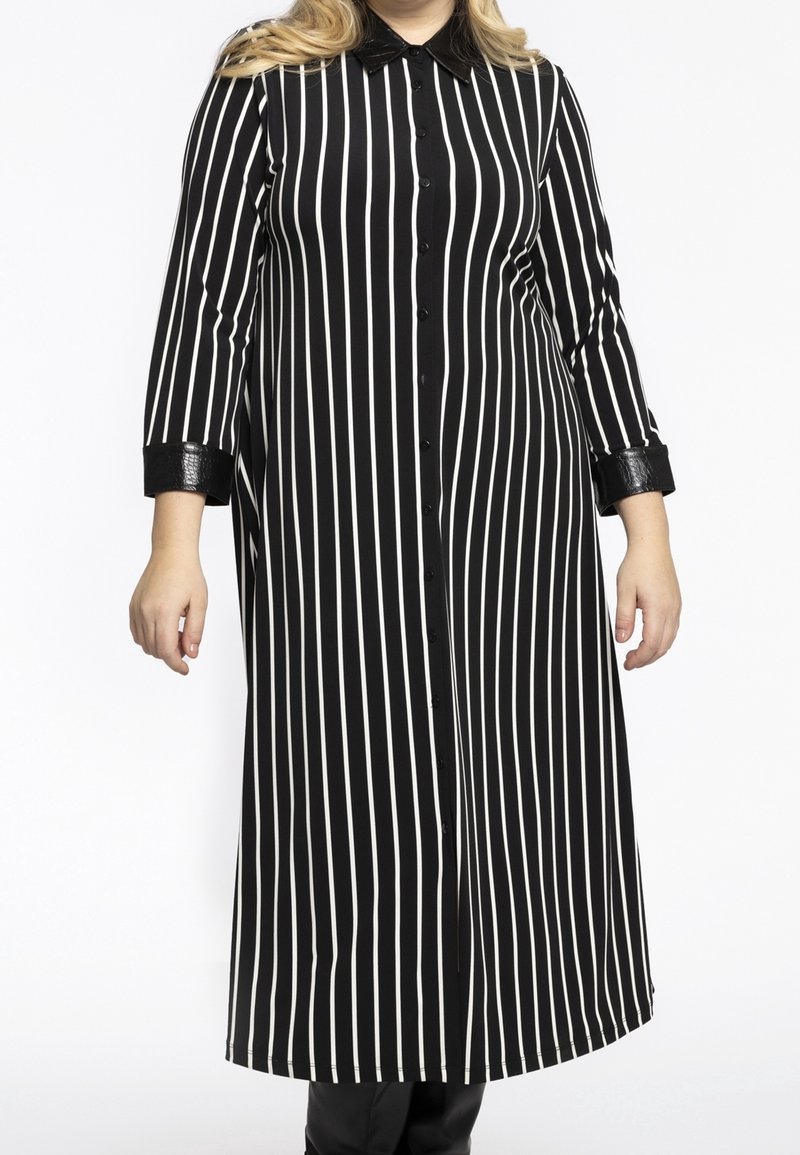 Femme portant une longue robe noire avec des rayures blanches verticales, un col noir brillant et des poignets noirs, debout sur un fond blanc.