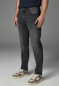 Schmal geschnittene, dunkelgraue Jeans mit leichtem Used-Look; verfügt über ein klassisches Fünf-Taschen-Design und einen Knopfverschluss. Getragen mit grauen Sneakers.