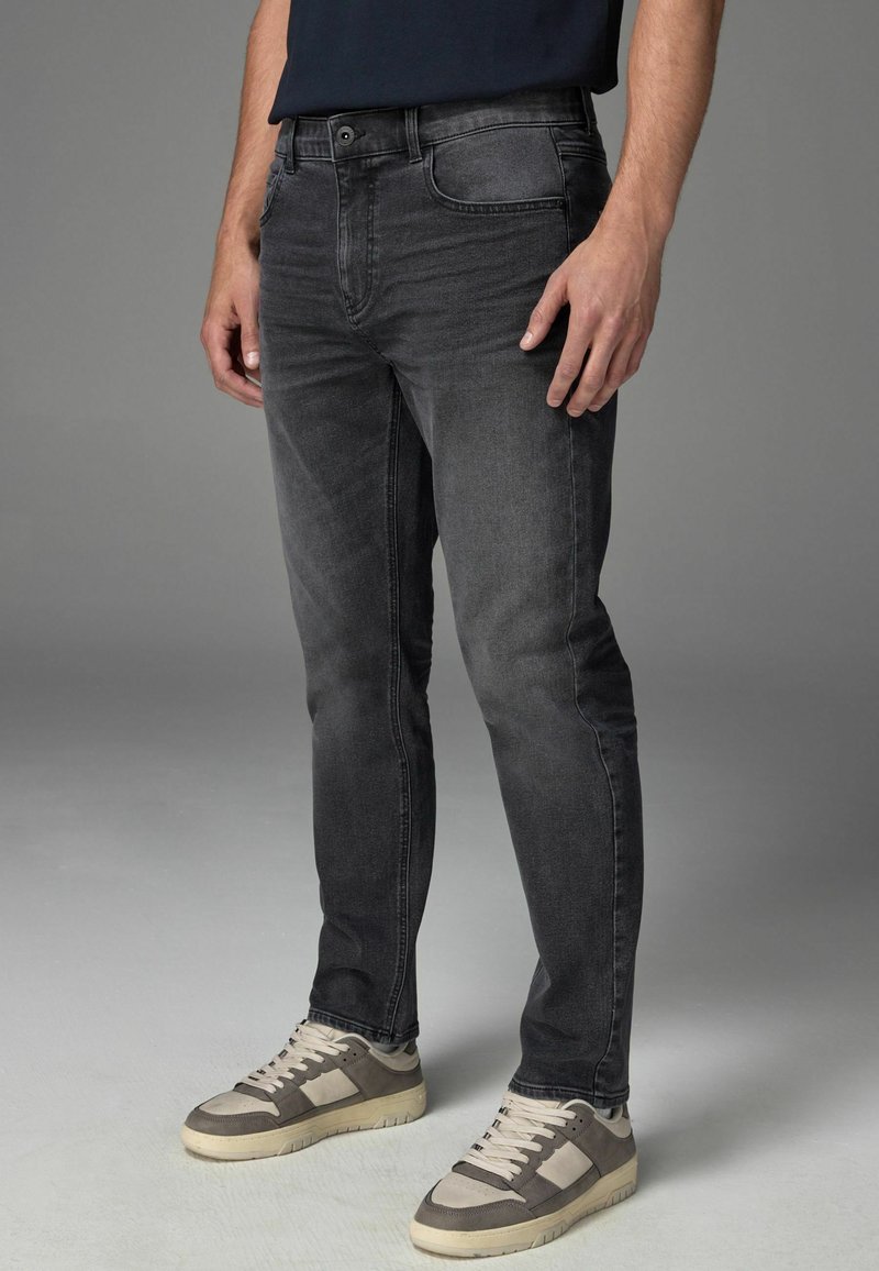 Schmal geschnittene, dunkelgraue Jeans mit leichtem Used-Look; verfügt über ein klassisches Fünf-Taschen-Design und einen Knopfverschluss. Getragen mit grauen Sneakers.