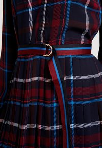 Zbliżenie na plisowaną sukienkę w granatowo-czerwono-niebiesko-białą kratkę, z pasującym paskiem w prążki z metalowym podwójnym zapięciem.