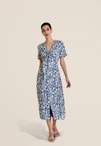 Robe chemise à motifs floraux bleus et blancs, longueur genoux, avec manches courtes et taille à nouer. Présente une fente devant pour faciliter les mouvements.