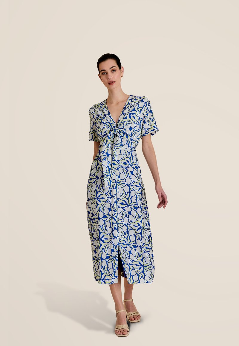 Robe chemise à motifs floraux bleus et blancs, longueur genoux, avec manches courtes et taille à nouer. Présente une fente devant pour faciliter les mouvements.