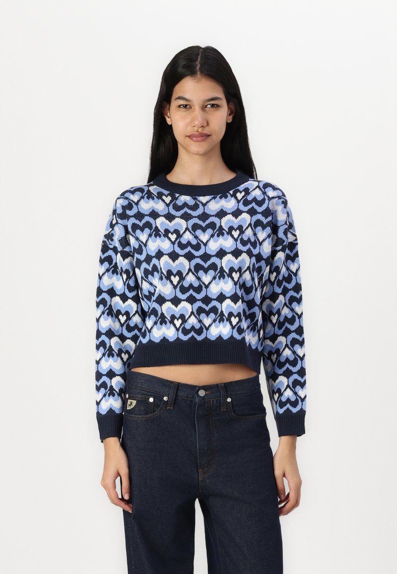 Blue cropped sweater featuring a heart pattern in varying shades with a ribbed neckline and cuffs, paired with dark blue jeans.