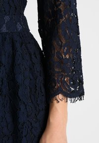 Granatowy rękaw sukienki z koronki z kwiatowymi wzorami, przezroczystą tkaniną i delikatnymi, frędzlowanymi brzegami na gołym ramieniu na jednolitym jasnym tle.