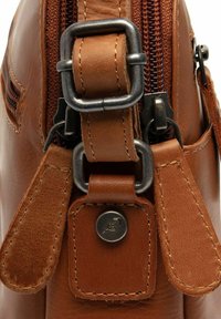 Dettaglio di una borsa in pelle marrone con un bottone a pressione metallico e zip, pelle testurizzata, cuciture decorative e un robusto attacco per la tracolla.