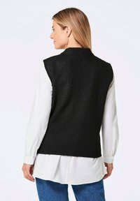 Ärmellose schwarze Strickweste mit strukturiertem Design und hohem Kragen. Getragen über einem weißen Hemd, kombiniert mit blauen Jeans.