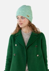 Jasnozielona dziana czapka z prążkowanym wzorem i metką z materiału. Noszona z ciemnozielonym podwójnym płaszczem i beżowym swetrem.