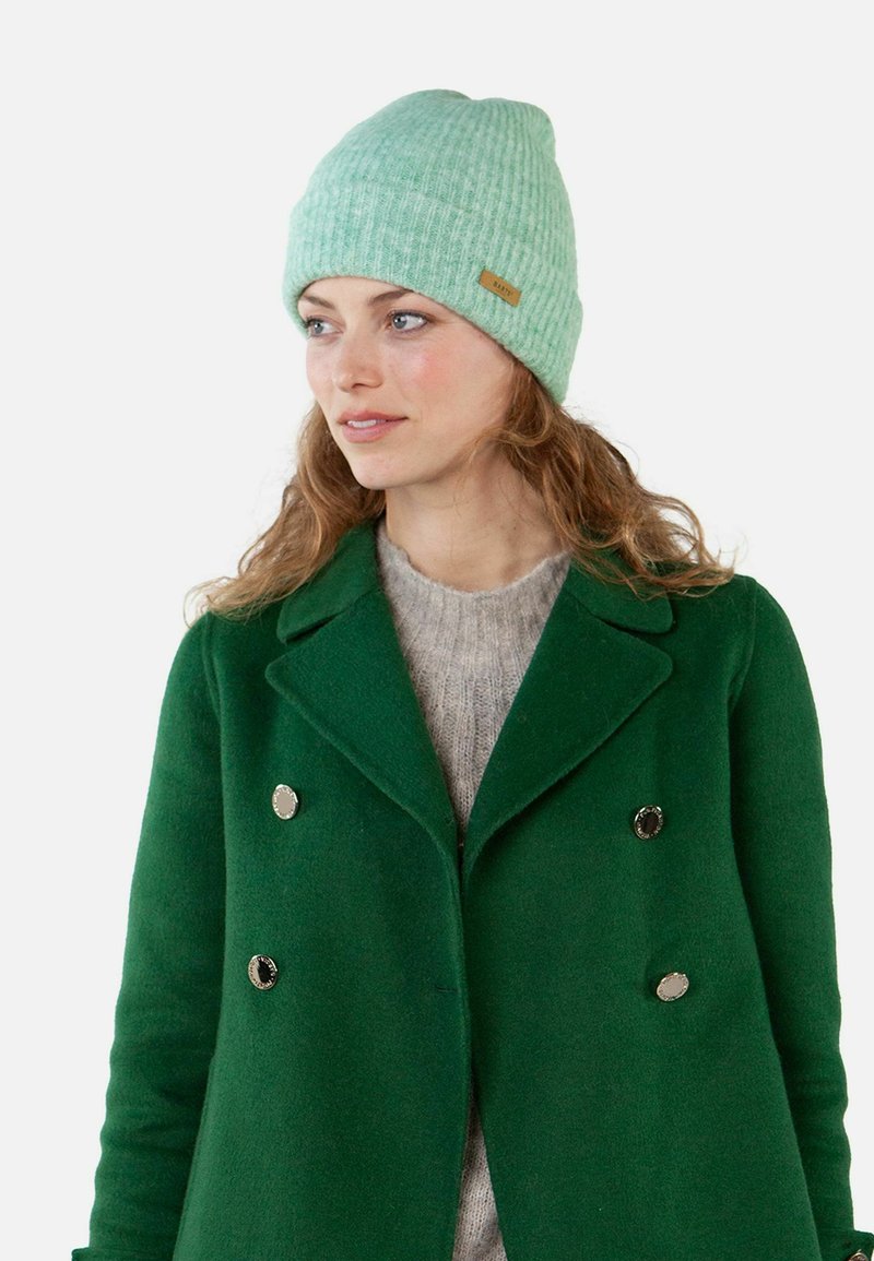 Jasnozielona dziana czapka z prążkowanym wzorem i metką z materiału. Noszona z ciemnozielonym podwójnym płaszczem i beżowym swetrem.