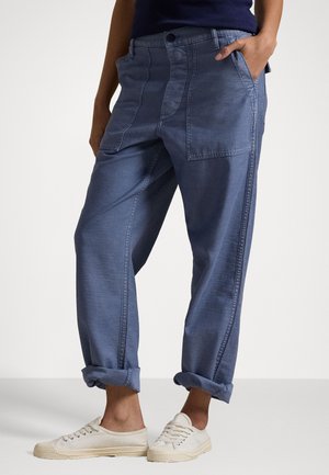 Baggy blue denim pants with rolled cuffs, featuring front pockets and a button closure. Worn with light-colored canvas sneakers.