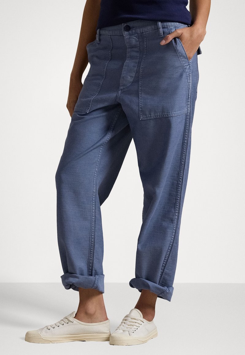 Pantaloni in denim blu larghi con risvolti, dotati di tasche frontali e chiusura a bottoni. Indossati con sneakers in tela di colore chiaro.
