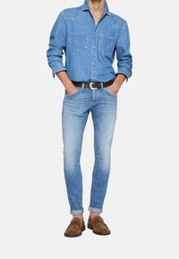 Jasnoniebieska koszula dżinsowa z dwiema kieszeniami na piersiach, w połączeniu z jasnymi, obcisłymi dżinsami i brązowymi zamszowymi loafersami, z podwiniętymi mankietami.