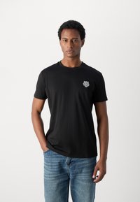 T-shirt en coton noir avec un col rond, des manches courtes, présentant un logo brodé détaillé sur la poitrine gauche. Porté avec un jean bleu.
