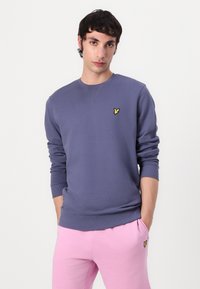 Sweatshirt violet foncé en matériau doux, avec un petit logo jaune sur la poitrine et des poignets et un ourlet côtelés. Associé à des joggers rose clair.