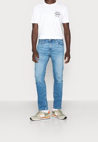Ljusblå denimjeans kombinerade med en vit T-shirt med svart grafik. Grå sneakers med inslag av orange och vitt.