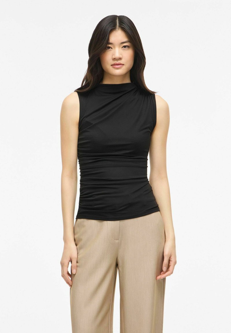Femme aux longs cheveux foncés portant un haut noir sans manches froncé et un pantalon beige à jambes larges, debout devant un fond uni.