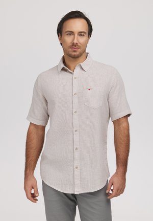 Mężczyzna ubrany w jasnobeżową koszulę na krótki rękaw z kieszenią na piersi i jasnoszare spodnie, stojący na jednolitym tle.