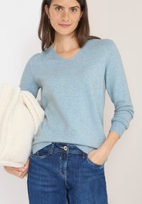 Jasnoniebieski sweter z dekoltem w serek, wykonany z miękkiego, teksturowanego materiału; ściągacze przy mankietach i u dołu; połączony z ciemnoniebieskimi dżinsami; trzyma w rękach kremowy polar.