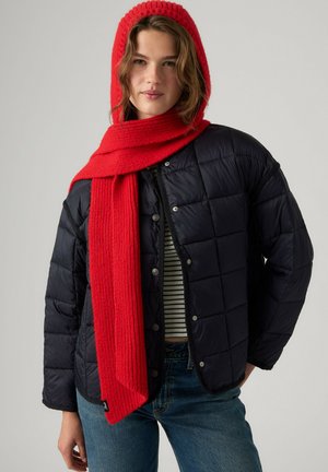 Woman wearing a red knit hooded scarf, black quilted jacket, striped top, and blue jeans against a plain gray background.