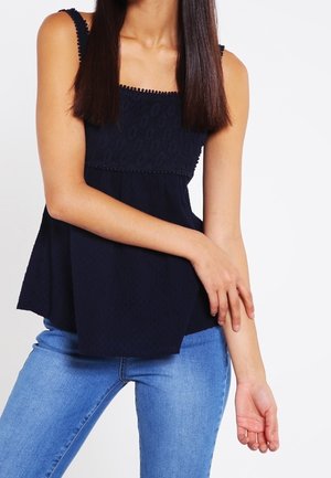 Vrouw die een donkerblauw mouwloos topje met kantdetails en lichtblauwe spijkerbroek draagt, poserend met één arm die de andere aanraakt.