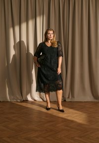 Femme en robe midi noire en dentelle et talons tient une pochette noire, debout sur un sol en bois avec des rideaux beiges en arrière-plan.