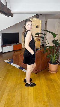 Rochie neagră fără mâneci din catifea, cu buzunare laterale, asortată cu pantofi loafer negri. Pardoseală din lemn și o plantă în ghiveci într-un decor interior modern.