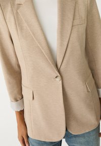 Blazer beige con un tessuto testurizzato, rever a bavero, chiusura con un bottone singolo e due tasche frontali. Sotto, top bianco a coste leggere.