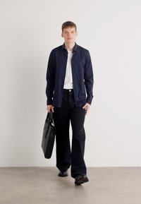 Cămașă albastră cu nasturi purtată peste un tricou alb, combinată cu pantaloni închiși la culoare și pantofi negri. Ținând o geantă tote neagră. Design minimalist.