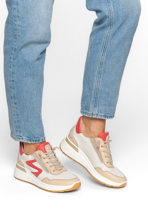 Person wearing light blue jeans and beige sneakers with red accents, standing with one foot slightly raised on a white background.