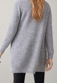 Frau mit langen Haaren, die eine hellgraue Strickjacke und schwarze Hosen trägt, von hinten gezeigt.