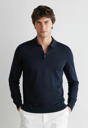 Mężczyzna o brązowych włosach, ubrany w granatową koszulkę polo z długim rękawem i białe spodnie, stoi z rękami w kieszeniach na jednolitym tle.