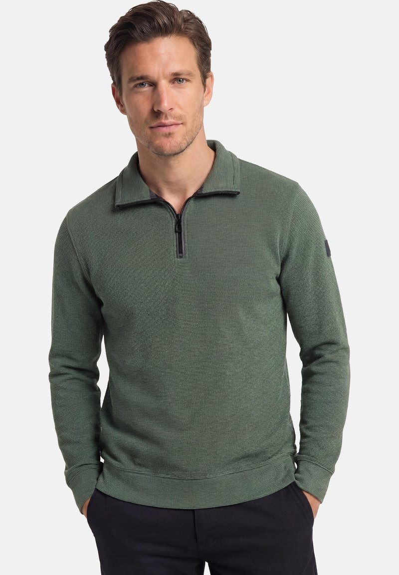 Uomo con capelli castani che indossa un maglione verde a mezza cerniera testurizzato e pantaloni scuri con le mani in tasca, in piedi davanti a uno sfondo semplice.