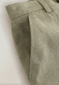 Gros plan sur un pantalon en tissu vert olive clair montrant la ceinture, les passants de ceinture, la poche latérale et les coutures détaillées.