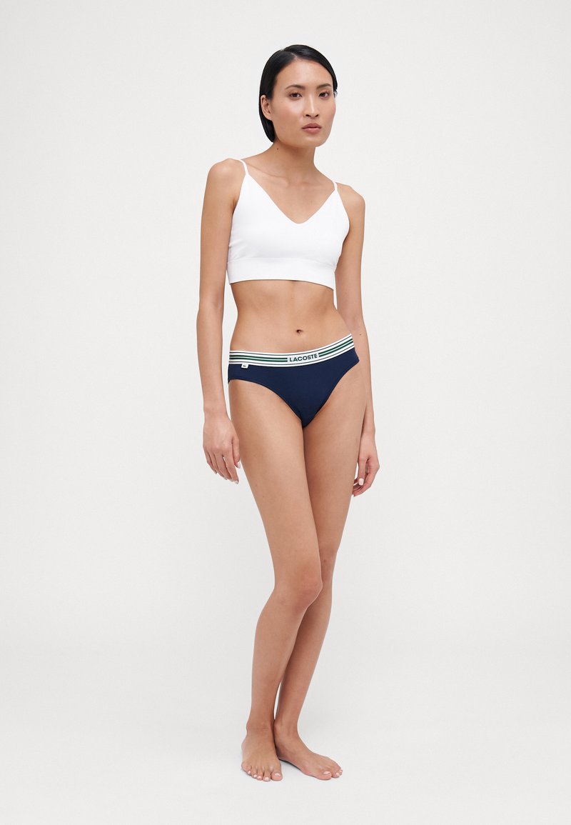 Bralette bianco in cotone con spalline sottili e scollatura a V. Slip bikini blu scuro con vita a righe in verde e bianco. Design minimalistico.