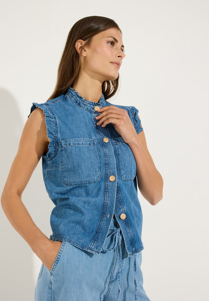 Vrouw draagt een mouwloos blauw denim gilet met knopen en een geplooide kraag, poseert met één hand bij de kraag en de andere in de zak.