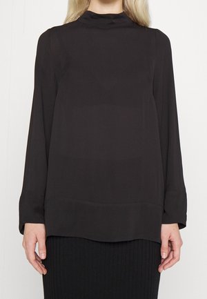 Nainen, jolla on pitkähihainen musta paita korkealla kauluksella, yhdistettynä resoripintaiseen mustaan hameeseen, yksiväristä vaaleaa taustaa vasten.