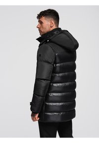 Hombre con cabello corto oscuro que lleva una chaqueta acolchada negra con capucha, de pie de perfil contra un fondo claro y liso.