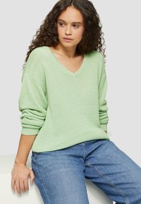 Hellegrüner, strukturierter Strickpullover mit weitem V-Ausschnitt und umgeschlagenen Ärmeln. Kombiniert mit blauen Jeans, sitzt vor einem neutralen Hintergrund.