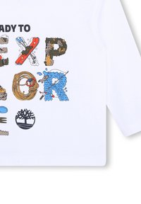 Witte T-shirt met kleurrijke grafische letters die "KLAAR OM TE ONTDEKKEN" spellen, vergezeld van natuurthema-illustraties, waaronder water, bomen en sportuitrusting.